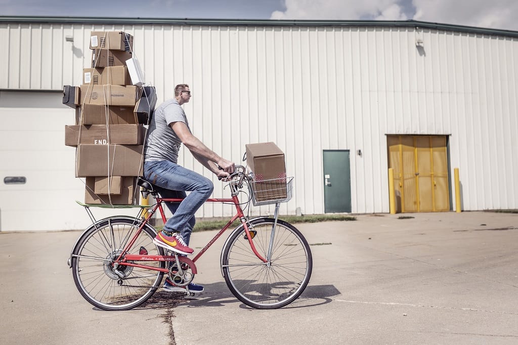 Man biking with many boxes attached to the back of the bike with a small head